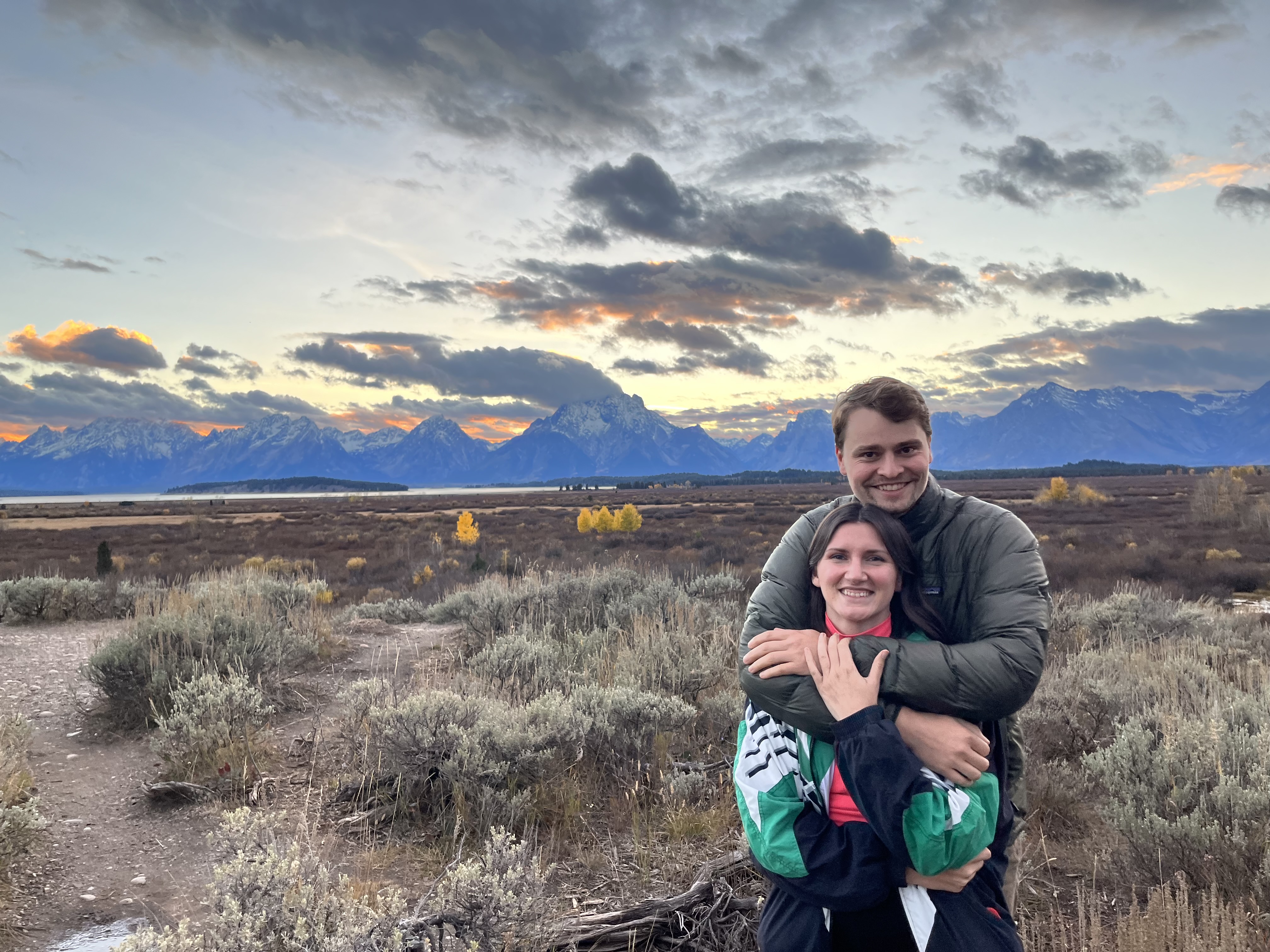 Connor and Jillian in front of a mountain range with the sun setting behind the mountains, making the sky orange